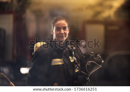 female firefighter portrait wearing full equipment and emergency rescue equipment. taking off oxygen mask after successful intervention. smoke and fire trucks in the background. 
