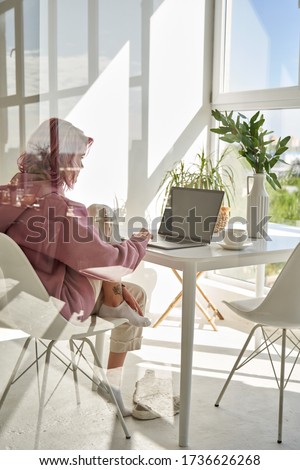 Young adult hipster teen girl school student with pink hair using laptop computer sitting at chair kitchen table behind glass working learning online at home office having morning breakfast. Vertical.
