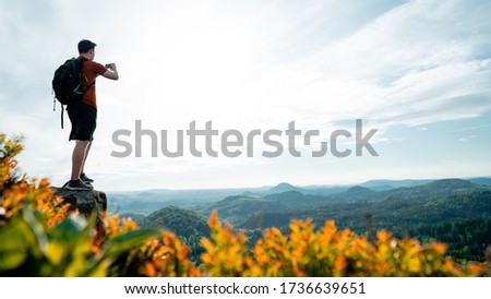 Hiker taking pictures on mobile in spring mountains. Scenic landscape of Lusatian Mountains, Czech Republic. Tourist on the rocky edge take phone pictures. Man is taking the landscape photos