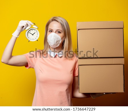 Blonde girl in face mask with delivery boxes and alarm clock on yellow background.