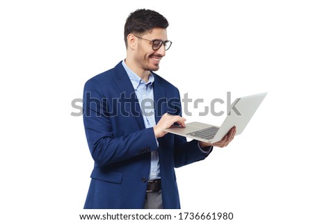 Smiling young man standing in blue suit and glasses, holding open laptop, watching funny content, feeling happy and relaxed, isolated on white background