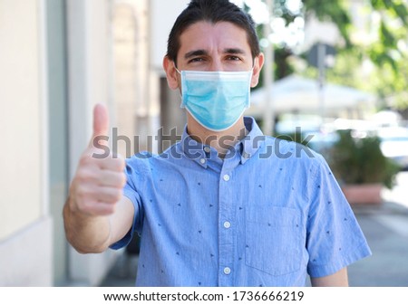 COVID-19 Optimistic young man wearing surgical mask on face during pandemic coronavirus disease showing thumbs up in city street Royalty-Free Stock Photo #1736666219