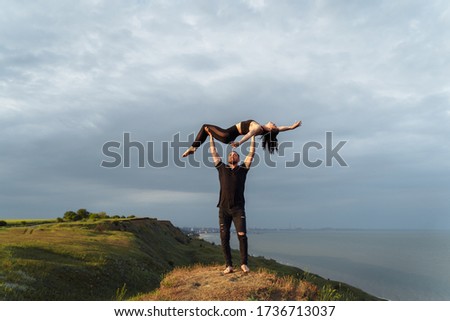 Full-length portrait. Attractive muscular, pumped-up young man is doing in acroyoga with his girlfriend. Instead of a bar, he lifts her up on outstretched arms and smiles. Standing on the edge of hill