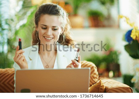 happy elegant female in white blouse and jacket with laptop and two lipsticks vlogging in the modern house in sunny day.
