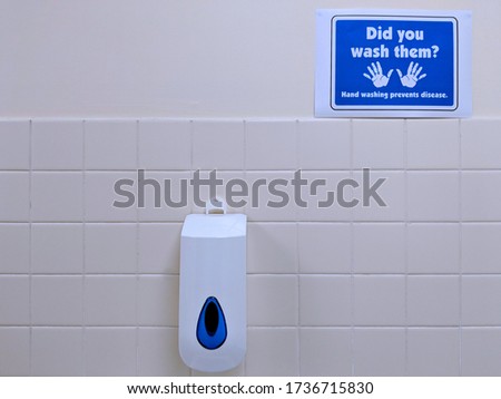 Blue and white "Did you wash them, Hand washing prevents disease" sign with a pair of hands icon and a liquid soap dispenser, on a magnolia, half tiled toilet wall in an office
