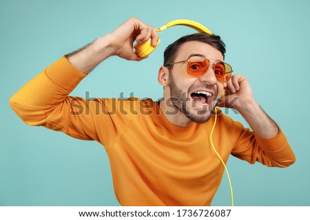Fashion bearded young man wearing sunglasses listening to music with yellow headphones on cyan background.