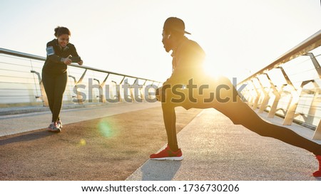 Happy african girl smiling and taking photo of man stretching on the bridge. He is warming up and smiling at camera. African couple running together in the morning. Sport. Training. Healthy lifestyle