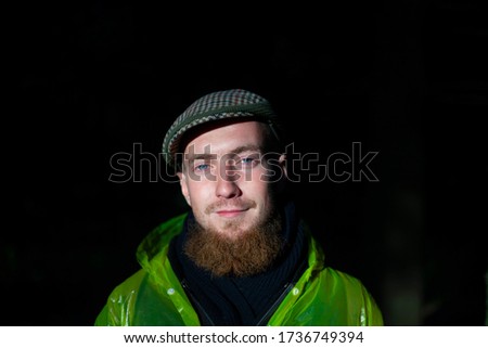 A guy with a long beard and an old-fashioned cap. Natural light when shooting. A lit face against a deep black background. Blue eyes in a man. Modern style of clothes. Beautiful portrait.