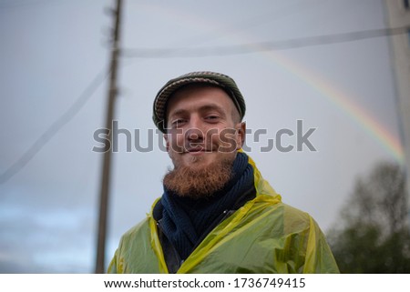 Portrait of a guy in a yellow rain jacket on a rainbow background. A young man with a big beard stands on the street after a summer rain. A man in an old-fashioned cap with blue eyes.