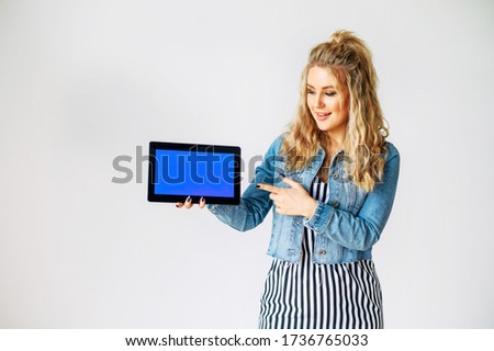 Look at this. Beautiful young woman with a tablet computer in hands stands on a white background. Woman points finger to blank tablet screen, empty space for inserting pictures, text
