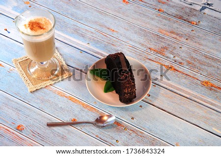 Ceramic plate with a piece of chocolate muffin and a tall glass with fresh cappuccino on a napkin. Close-up.