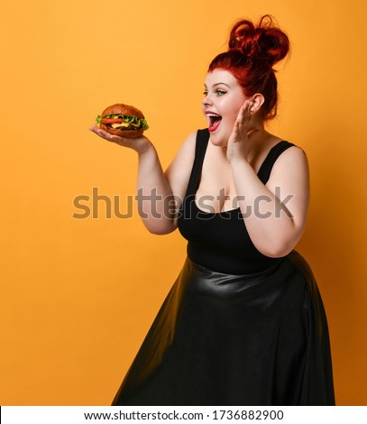 Happy screaming plus-size woman looks at burger cheeseburger sandwich with beef she holds on her open palm on yellow background with copy space. Healthy eating diet fast food concept