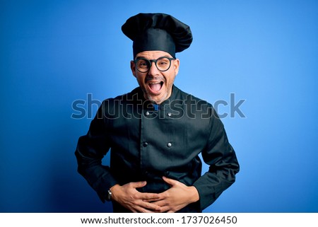 Young handsome chef man wearing cooker uniform and hat over isolated blue background smiling and laughing hard out loud because funny crazy joke with hands on body.
