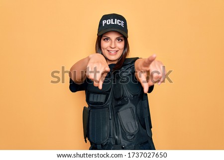 Young beautiful brunette policewoman wearing police uniform bulletproof and cap pointing to you and the camera with fingers, smiling positive and cheerful