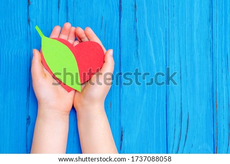 Top view of green leaf with red heart in hands of child on wooden blue background. Copy space. Concept of environmental protection and Earth day.