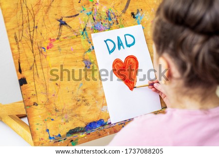 Child's hand drawing red heart with word Dad greeting card on white paper on an easel. Happy Father's Day concept.