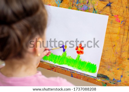 Hand of little girl drawing flowers on green grass with brush by watercolors on white paper on an easel.