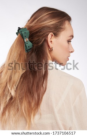 A back turned blonde girl with golden earrings is wearing a white blouse. The lady's hair is fixed with a hair clip decorated with a green bow with polka dot print, a golden buckle and a silk ribbon.  Royalty-Free Stock Photo #1737106517