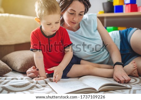 Young mother with dreadlocks on her head and her little son are reading a book together. Concept of happy communication with your child, the joy of being with your family at home.