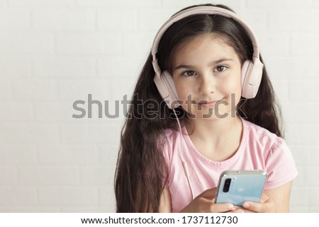Online talking. Cute little girl in headphones is using a smartphone, looking at camera and smiling on light background. Listening music. 