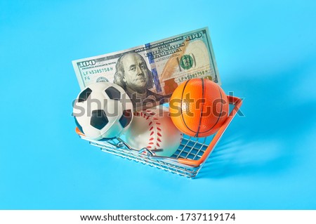 Different balls and banknote of 100 dollars in metal market basket on blue background. Purchasing sport accessories. Concept of corruption in sport
