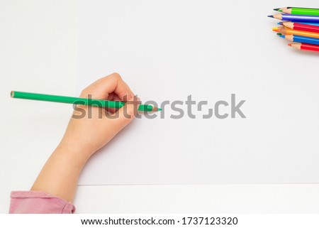 Top view of child's left hand drawing at blank white paper with a green pencil. Copy space for text. Mockup.