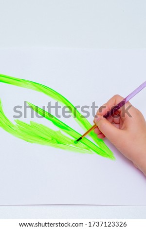 Child's hand drawing green leaf by watercolors on white sheet of paper. Earth day concept. Top view.
