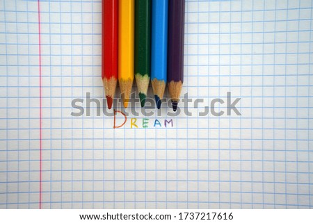 The inscription DREAM written in colored pencils, next to a checkered sheet of paper lay pencils                  