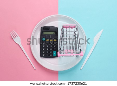 Supermarket trolley and calculator in plastic plate with fork and knife on pink blue pastel background. Food purchase. Top view