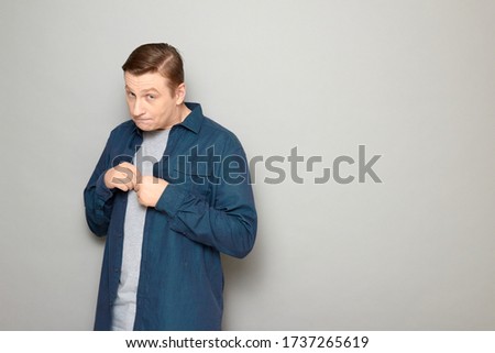 Studio portrait of shy blond mature man wearing casual blue shirt, looking naive and timid, apologizing, asking for forgiveness, feeling guilty, standing against gray background, copy space on right Royalty-Free Stock Photo #1737265619