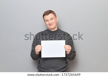 Studio portrait of happy blond mature man wearing jumper, smiling cheerfully, holding white blank paper sheet with place for your text in hands, standing over gray background