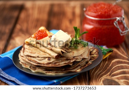  A stack of freshly baked hot pancakes with a piece of butter on a dark plate on a folded towel with a glass jar filled with red caviar. Russian traditions, carnival. Top view, place for text.         Royalty-Free Stock Photo #1737273653