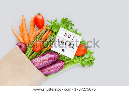 Fresh organic vegetables in eco craft paper shopping bag with text Buy Local on lightbox flat lay, top view with copy space on gray background. Sustainable lifestyle. Zero waste, plastic free concept.