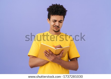 Photo of thinking african american man grimacing and making notes in planner isolated over purple background