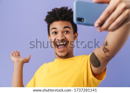 Photo of cheerful african american man taking selfie on smartphone and holding copyspace isolated over purple background