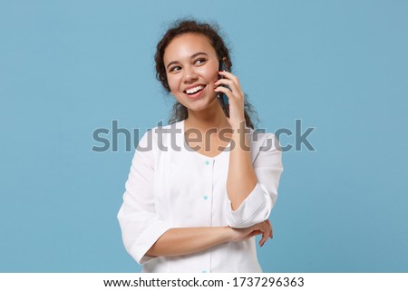 Cheerful african american doctor woman isolated on blue wall background. Female doctor in white medical gown talking on mobile phone. Healthcare personnel health medicine concept. Mock up copy space