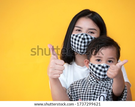 Asian mother and son are showing thumb up wearing mask trying to protect from coronavirus epidemic over yellow background.