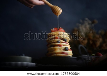 Pouring honey on stack of tasty pancakes with fresh blueberries, strawberries and oranges.