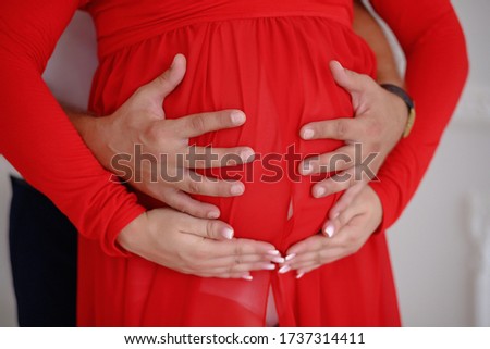 Man and woman hold by hands the pregnant belly of his wife in a red dress. Loving couple awaiting the birth of a baby. Close-up photo of hands and abdomen without faces