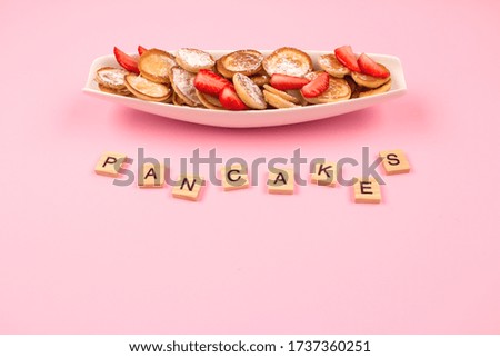 Flat lay minimal composition with mini pancakes cereal with strawberries on white plate and wooden letters on pink background with copy space for recipe or your text. Breakfast concept, trendy food