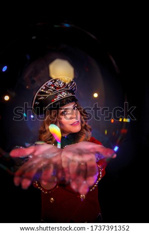 Girl in stage costume and cap on her head. Female magician, an illusionist in theatrical clothes, holds soap bubble into camera on black background. Concept of theatrical performance