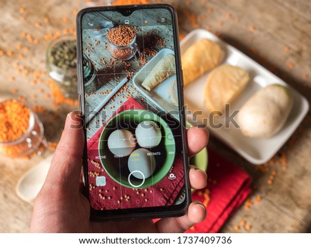 Taking a photo with the mobile phone of a presentation of orange lentils inside glass of jars and sunflower seeds on a top of wooden table with eggs and other vegetables seen from above.