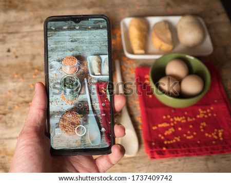 Taking a photo with the mobile phone of a presentation of orange lentils inside glass of jars and sunflower seeds on a top of wooden table with eggs and other vegetables seen from above.