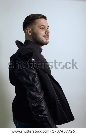 Young Bearded Dark Haired Man In Black Stylish Shirt, Jacket On Gray Background, Serious Male Sheds Outerwear, Undresses. The Concept Of Posing Models