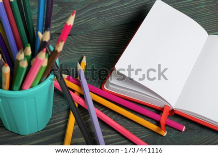 Pencil filled with colored pencils. In the form of a garbage container. Nearby is an open notebook and several pencils. Against the background of brushed pine boards painted in black and green.