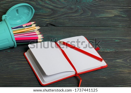 A pencil in the form of a garbage container filled with colored pencils lies on its side. Near an open notebook. Against the background of brushed pine boards painted in black and green.