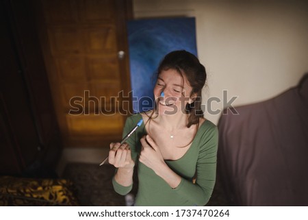 Brunette girl with closed eyes and freckles on her cheeks escape isolation in her workshop, drawing a blue picture, smiling, having fun 