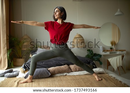 People, activity, health and vitality concept. Stylish barefoot young woman exercising at home, doing vinyasa flow yoga in her bedroom, standing on carpet in virabhadrasana or warrior II pose Royalty-Free Stock Photo #1737493166