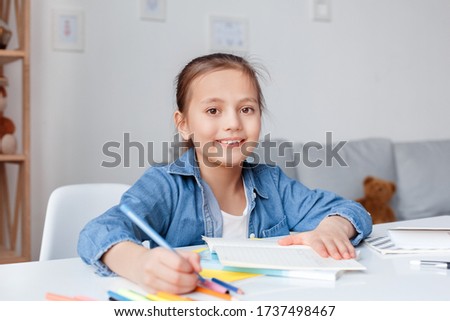 Little cute pretty girl doing lessons writing in exercise book after school. Prepares homework assignment, concentrated focus for teach read study. Looks into camera smiley. Face portrait close up.