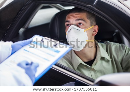 Male driver wearing protective N95 face mask sitting by left drive wheel in UK drive-thru COVID-19 test centre,answering health check up questions,medical worker ticking off symptoms on clipboard form Royalty-Free Stock Photo #1737555011